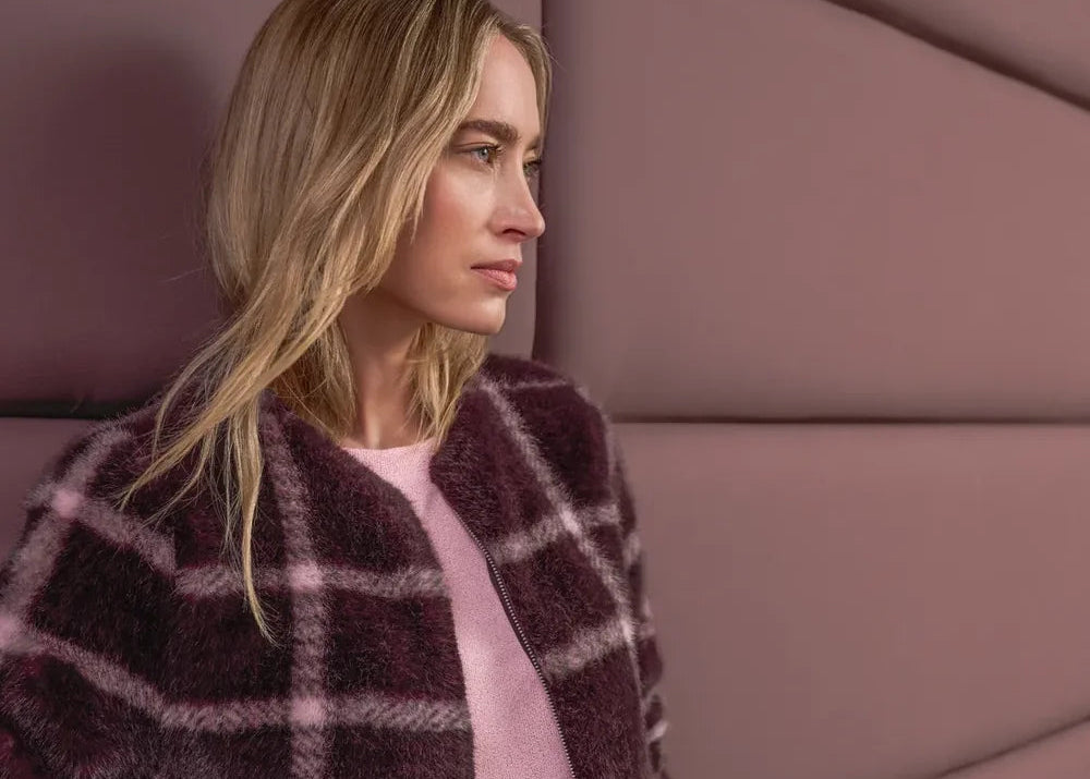 A woman wearing a fuzzy dark plum plaid cardigan over a light pink top and blue jeans, standing against a mauve padded wall.