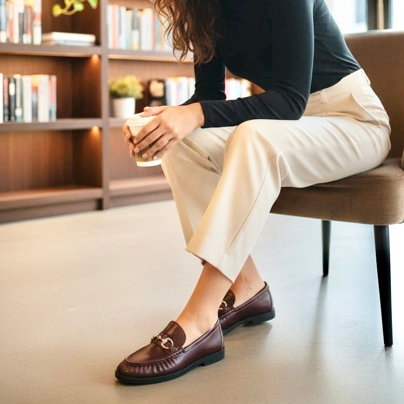 A woman sits on a brown upholstered chair in a modern indoor setting with bookshelves in the background. She is wearing a black long-sleeve top, cream-colored pants, and dark brown leather loafers with gold hardware.
