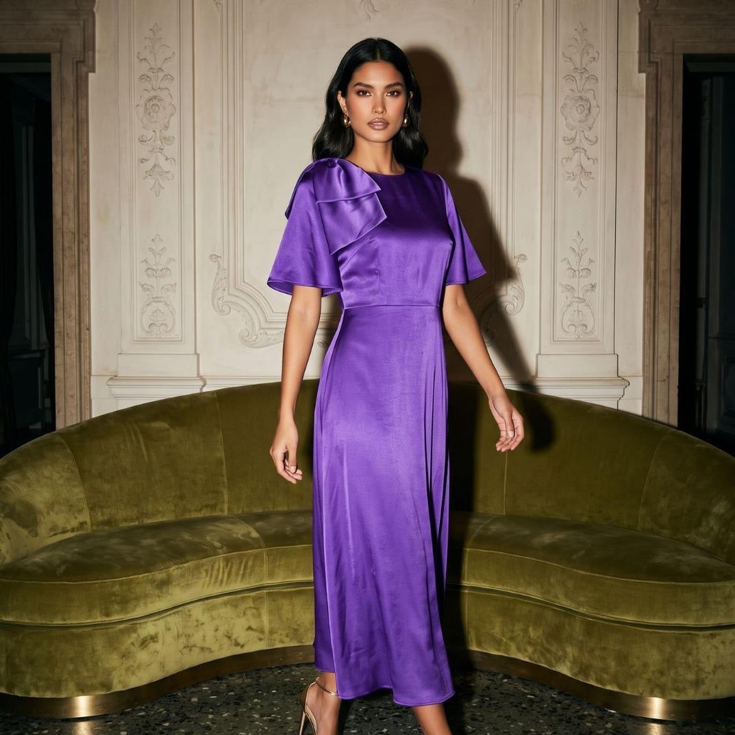 A woman models a purple satin midi dress with a ruffled shoulder detail in an elegant room with a green velvet curved sofa and ornate wall molding.